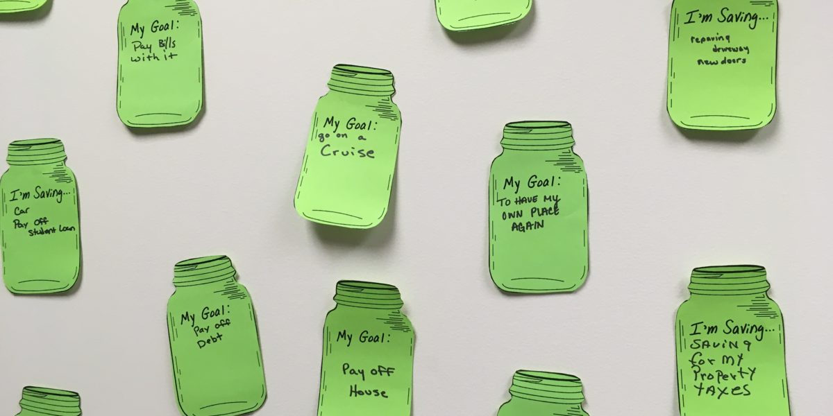 Green paper mason jars with savings goals written on them taped to a white wall.