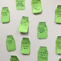Green paper mason jars with savings goals written on them taped to a white wall.