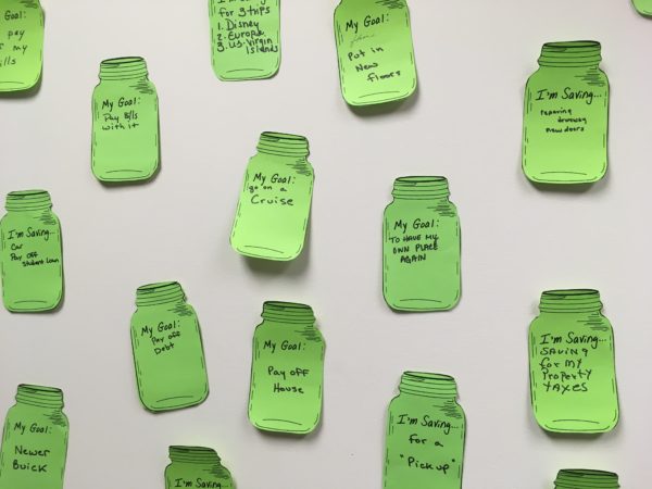 Green paper mason jars with savings goals written on them taped to a white wall.