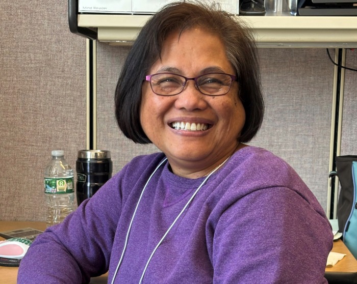 Francia has shoulder length hair and glasses and is smiling at the camera and wearing a purple sweater